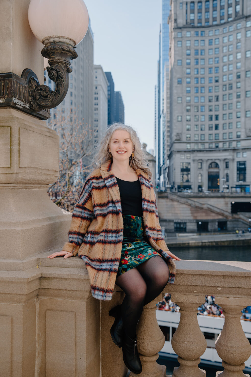 Photo of Ana Pruteanu sitting on a bridge downtown Chicago, skyline in background. Ana is smiling directly at the camera. 