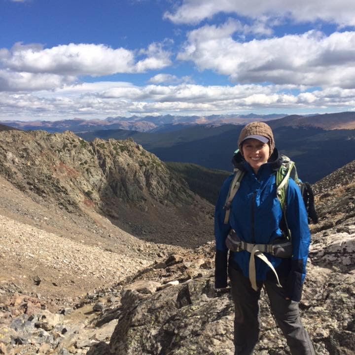 Ana backpacking in Colorado, mountains in the background. Ana is wearing a blue jacket and backpack.