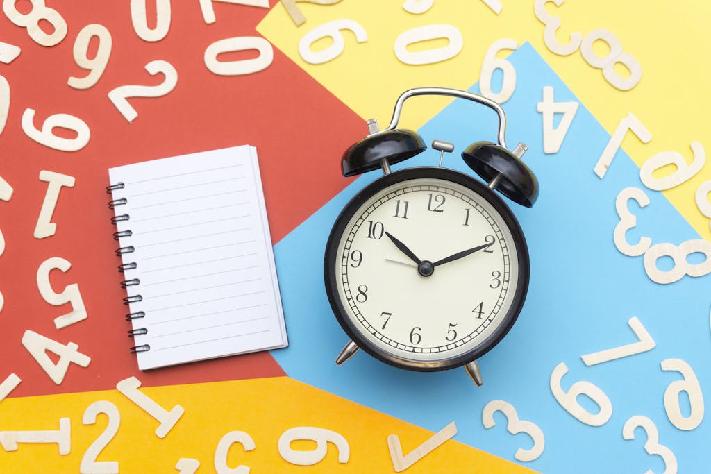 A flat lay of a classic alarm clock with a blank notebook on a colorful background surrounded by numbers.