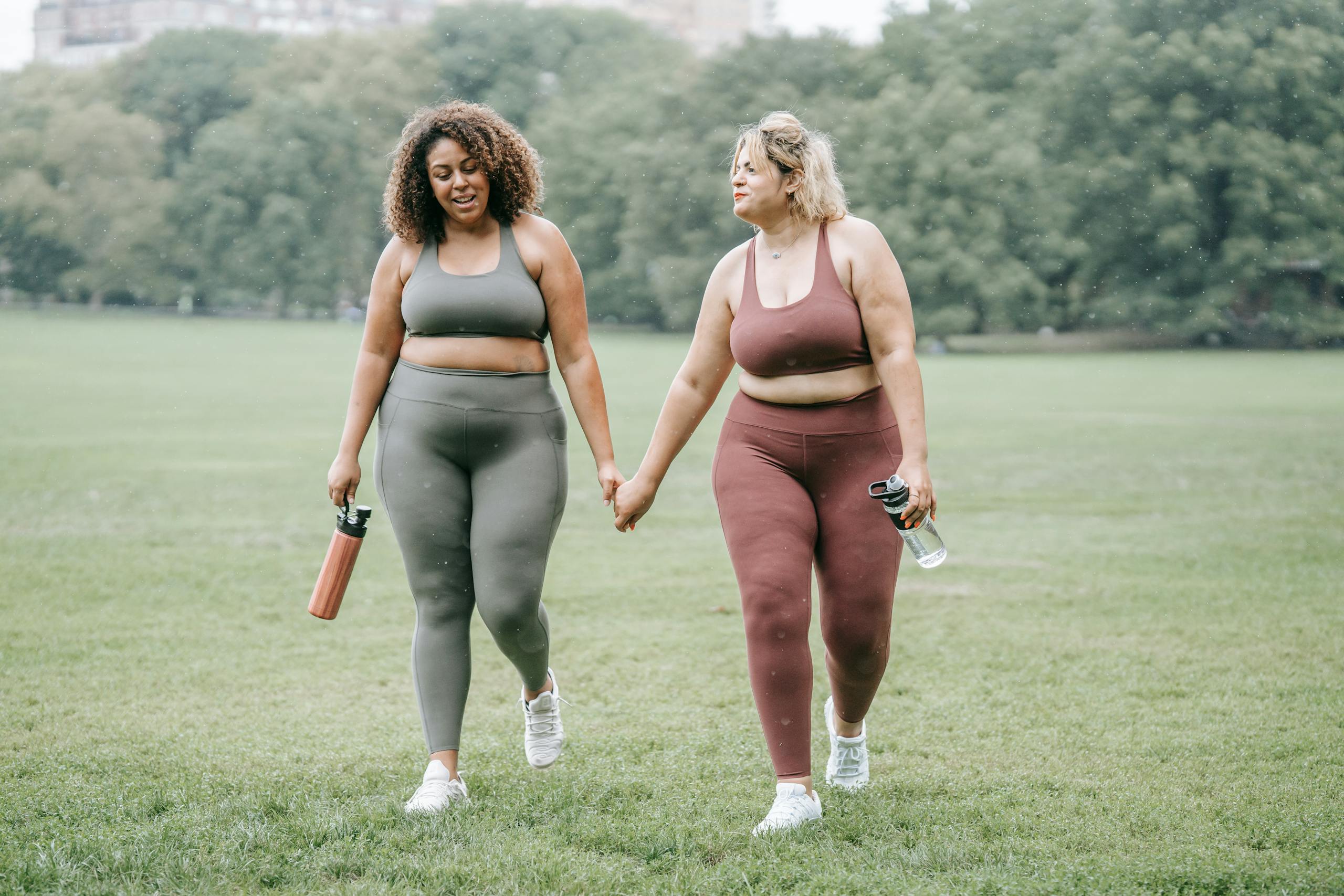 Two plus-size women in activewear holding hands while walking in the park